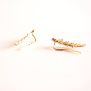 May include: A pair of gold leaf ear cuffs. The cuffs are made of a thin wire that is shaped into a leaf design. The cuffs are designed to be worn on the upper part of the ear.