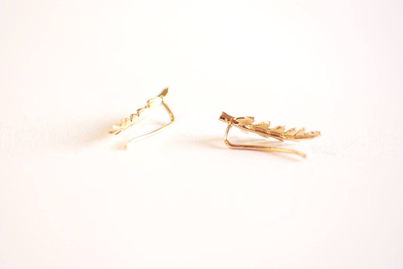 May include: A pair of gold leaf ear cuffs. The cuffs are made of a thin wire that is shaped into a leaf design. The cuffs are designed to be worn on the upper part of the ear.