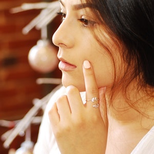 May include: A close-up of a woman's face, with her finger touching her cheek. She is wearing a silver ring with a leaf design on her ring finger.
