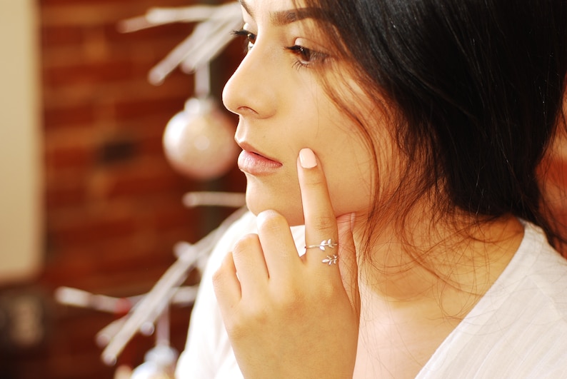 May include: A close-up of a woman's face, with her finger touching her cheek. She is wearing a silver ring with a leaf design on her ring finger.