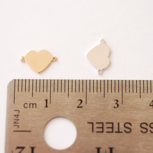 May include: Two heart-shaped metal charms, one gold and one silver, are shown next to a ruler. The charms are approximately 1 inch in size.