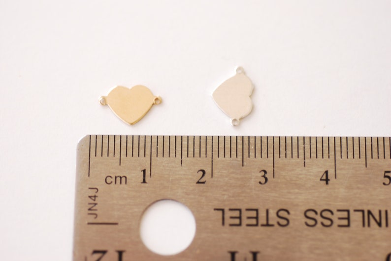 May include: Two heart-shaped metal charms, one gold and one silver, are shown next to a ruler. The charms are approximately 1 inch in size.