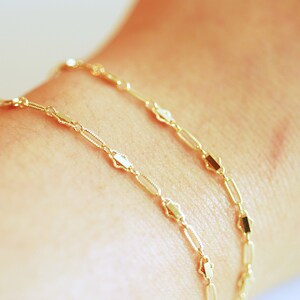 May include: Two gold chain bracelets with a delicate, linked design. The chains feature small, rectangular-shaped links.