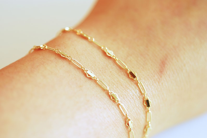 May include: Two gold chain bracelets with a delicate, linked design. The chains feature small, rectangular-shaped links.