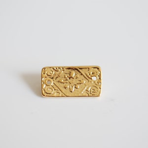 May include: Gold-coloured metal charm shaped like a rectangular biscuit with a raised design.