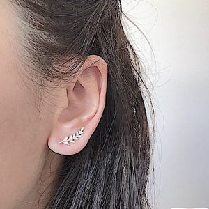May include: A close-up of a person's ear with a small, delicate silver leaf earring. The earring is made of a thin, delicate wire with small, clear stones set into the leaf shape. The earring is worn in the upper lobe of the ear.