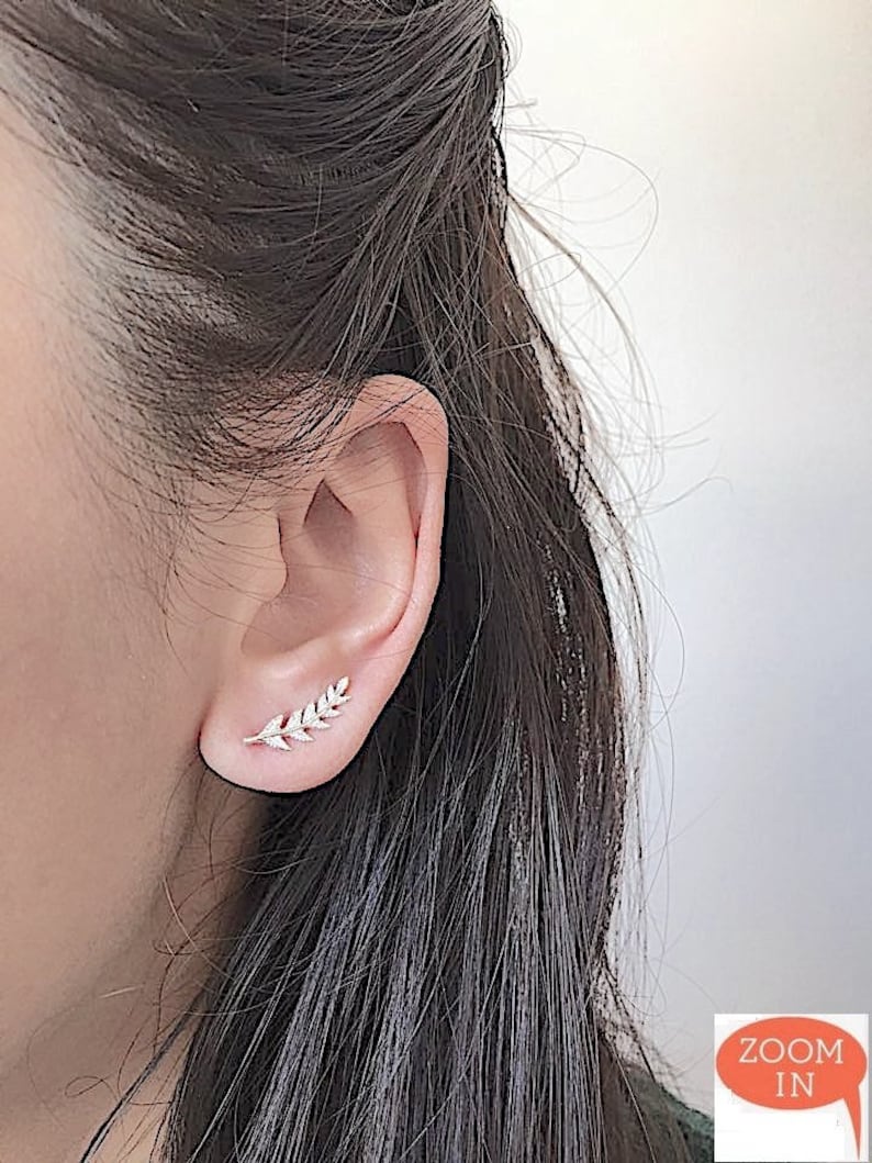 May include: A close-up of a person's ear with a small, delicate silver leaf earring. The earring is made of a thin, delicate wire with small, clear stones set into the leaf shape. The earring is worn in the upper lobe of the ear.