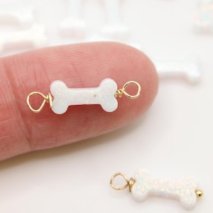 May include: A white, bone-shaped charm with an iridescent finish, featuring gold-toned wire loops on both ends. The charm is small and designed for jewelry making, with a smooth, polished surface that reflects light with subtle colorful glints.