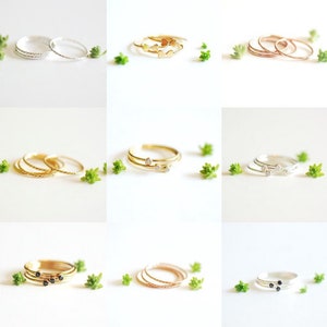 May include: Set of delicate gold, silver, and rose gold stacking rings with various designs, including hearts, diamonds, and black stones. The rings are displayed on a white background with small green plants.