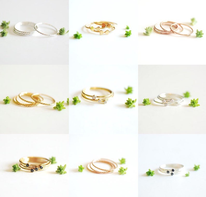 May include: Set of delicate gold, silver, and rose gold stacking rings with various designs, including hearts, diamonds, and black stones. The rings are displayed on a white background with small green plants.