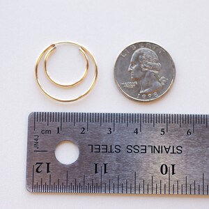 May include: Two gold hoop earrings, one slightly larger than the other, are shown next to a US quarter for size comparison. A ruler is also included in the image.