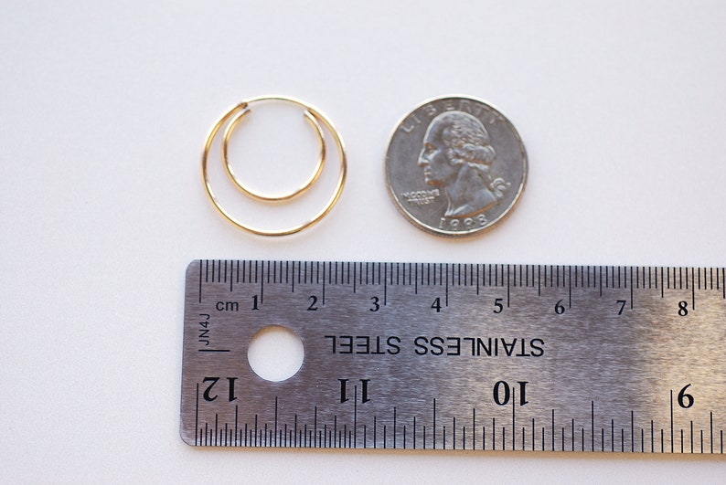 May include: Two gold hoop earrings, one slightly larger than the other, are shown next to a US quarter for size comparison. A ruler is also included in the image.