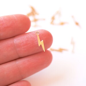 May include: A gold-colored lightning bolt charm with a small loop at the top. The charm is held in a hand against a white background.