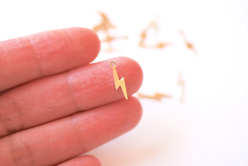 May include: A gold-colored lightning bolt charm with a small loop at the top. The charm is held in a hand against a white background.