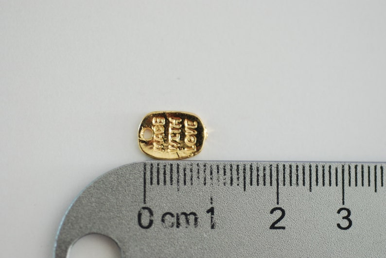 May include: Gold-coloured metal tag with the words "Made with Love" engraved on it. The tag is approximately 1 cm long and is next to a ruler.