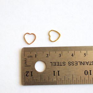 Puede incluir: Dos anillos de salto en forma de coraz&oacute;n, uno dorado y otro de cobre, se muestran junto a una regla. El anillo de salto dorado tiene un di&aacute;metro de 1 cm. El anillo de salto de cobre tiene un di&aacute;metro de 1,2 cm.