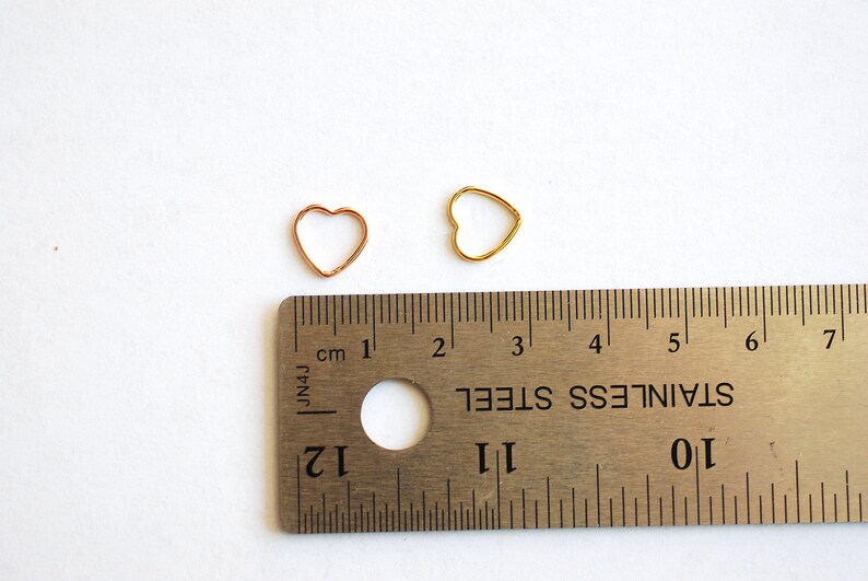 Puede incluir: Dos anillos de salto en forma de coraz&oacute;n, uno dorado y otro de cobre, se muestran junto a una regla. El anillo de salto dorado tiene un di&aacute;metro de 1 cm. El anillo de salto de cobre tiene un di&aacute;metro de 1,2 cm.