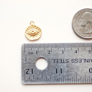 Puede incluir: Encanto dorado con un dise&ntilde;o de ojo detallado, que mide aproximadamente 1 cm de di&aacute;metro. El encanto se muestra junto a una moneda de 25 centavos de d&oacute;lar estadounidense para comparar el tama&ntilde;o.