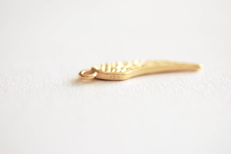 May include: A gold-colored wing charm with a textured surface. The charm has a loop for attaching to a necklace or bracelet. The charm is marked with the number "925".