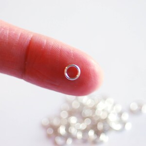 May include: A close-up of a silver jump ring held between a finger and thumb. The ring is small and round, with a smooth surface. A pile of similar jump rings is visible in the background.