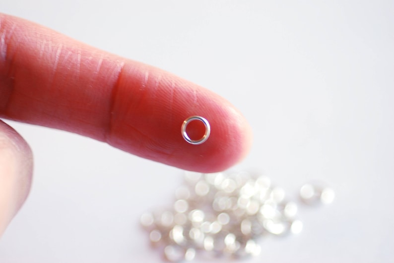 May include: A close-up of a silver jump ring held between a finger and thumb. The ring is small and round, with a smooth surface. A pile of similar jump rings is visible in the background.
