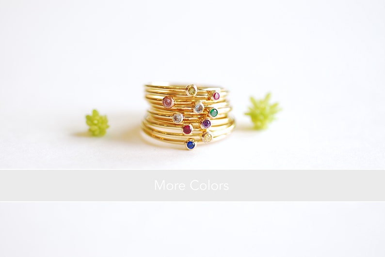 May include: A stack of gold rings with small colorful gemstones. The rings are stacked on top of each other and the gemstones are visible. The text "More Colors" is below the rings.