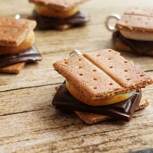 以下が含まれることがあります： グラハムクラッカー、マシュマロ、チョコレートスクエアで構成された、いくつかのミニチュアのS'moreのクローズアップ。S'moreは木製の表面に配置され、食品の詳細が強調されています。S'moreは薄茶色です。