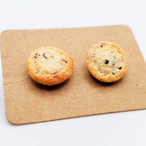 May include: Two miniature chocolate chip cookies on a brown paper card. The cookies have a golden-brown crust and a creamy, speckled interior with visible chocolate chips. The card is a light brown color.
