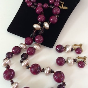 May include: A vintage jewelry set featuring a necklace and matching clip-on earrings. The necklace and earrings showcase large, round, deep burgundy beads interspersed with pearlescent and faceted dark beads. The set is displayed on a black velvet surface.