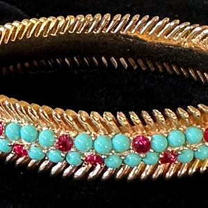 Op de afbeelding: Een gouden armband met een chevron-ontwerp en turquoise en rode stenen.