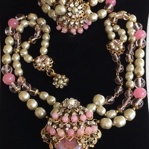 May include: A three-strand pearl necklace with pink glass beads and a large pink glass pendant with a gold filigree design and rhinestones. The necklace is paired with a matching bracelet.