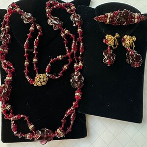 May include: A set of red glass jewellery featuring a long necklace, a brooch, and a pair of earrings. The necklace is made of two strands of red glass beads, some of which are shaped like roses. The brooch is a simple design with red glass beads and a gold-toned clasp. The earrings are drop earrings with red glass beads and gold-toned clasps.