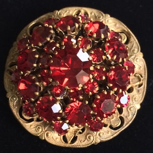 May include: A round, gold-toned brooch with a cluster of red rhinestones in the center. The brooch has a decorative, filigree design around the edge.