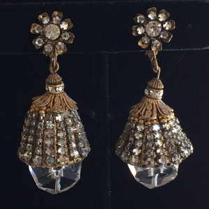 Puede incluir: Un par de pendientes colgantes de tono dorado con acentos de cristal transparente en forma de lágrima. Los pendientes presentan un racimo de diamantes de imitación en la parte superior y un racimo más pequeño de diamantes de imitación en la lágrima.