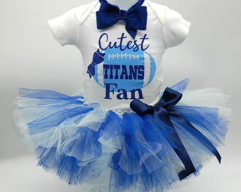 May include: A white baby onesie with blue and white football design and the text "Cutest Titans Fan". The onesie is paired with a blue tulle tutu and a navy blue bow tie.