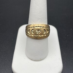 Może przedstawiać: Złoty pierścionek z szeroką obrączką. Pierścionek ma powtarzający się wz&oacute;r gwiazd i wycięć w kształcie liści wok&oacute;ł obrączki. Pierścionek jest prezentowany na białym, palcowatym stojaku na czarnym tle.