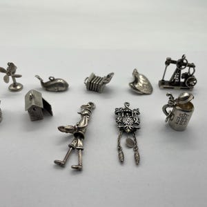 May include: A collection of ten silver-toned metal charms including a fan, a whale, an accordion, a leaf, an oil derrick, a set of teeth, a small house, a figure, a cuckoo clock, and a lidded beer stein with an HB logo and crown engraving.