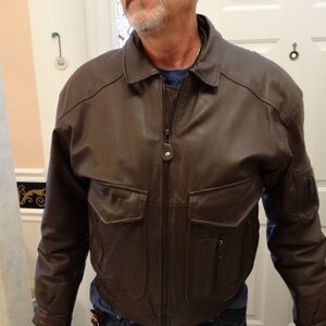 May include: A brown leather jacket with a zipper closure and two large pockets on the front. The jacket has a stand-up collar and a zippered pocket on the left sleeve.
