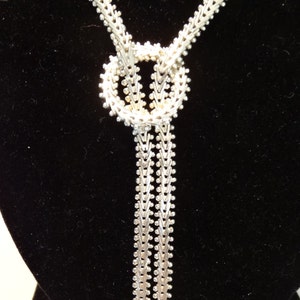 May include: A silver chain necklace with a knot design and two long strands. The necklace is displayed on a black velvet stand.