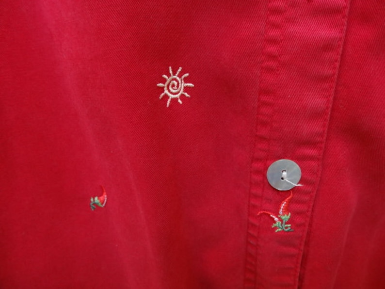 May include: Red cotton shirt with embroidered sun and chili pepper designs. The shirt has a button closure and a single button on the front.