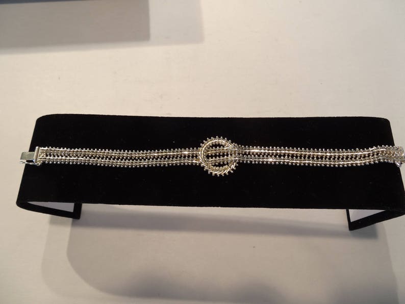 May include: A silver bracelet with a circular design in the center. The bracelet is made of multiple strands of small beads.