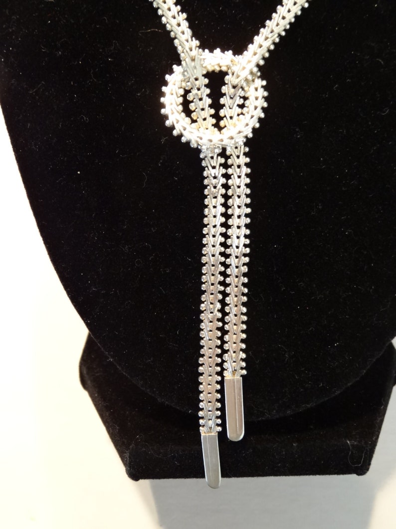 May include: A silver chain necklace with a knot design and two long strands. The necklace is displayed on a black velvet stand.