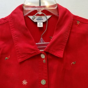 May include: Red button-down shirt with embroidered suns and peppers. The shirt has a collar and a button closure. The label reads "AKS Specialty XL".