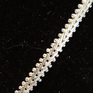 May include: A silver chain necklace with a geometric pattern. The chain is made of small, interconnected links that form a repeating pattern of triangles.