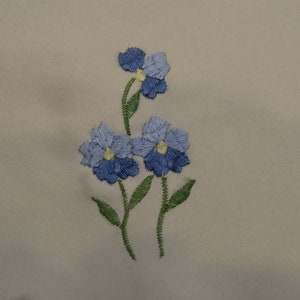 May include: Embroidered design of three blue flowers with yellow centers on a beige fabric background.