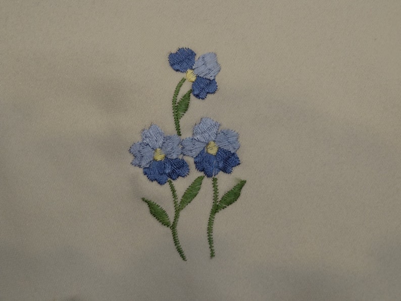 May include: Embroidered design of three blue flowers with yellow centers on a beige fabric background.