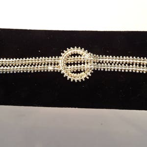 May include: A silver bracelet with a circular design in the center. The bracelet is made of multiple strands of small beads.