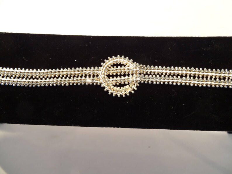 May include: A silver bracelet with a circular design in the center. The bracelet is made of multiple strands of small beads.
