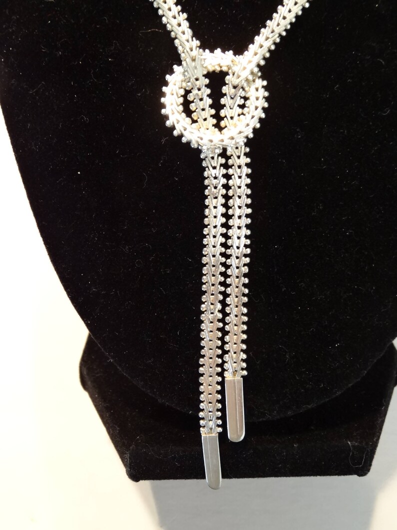 May include: A silver chain necklace with a knot design and two long strands. The necklace is displayed on a black velvet stand.