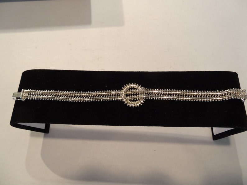 May include: A silver bracelet with a circular design in the center. The bracelet is made of multiple strands of small beads.